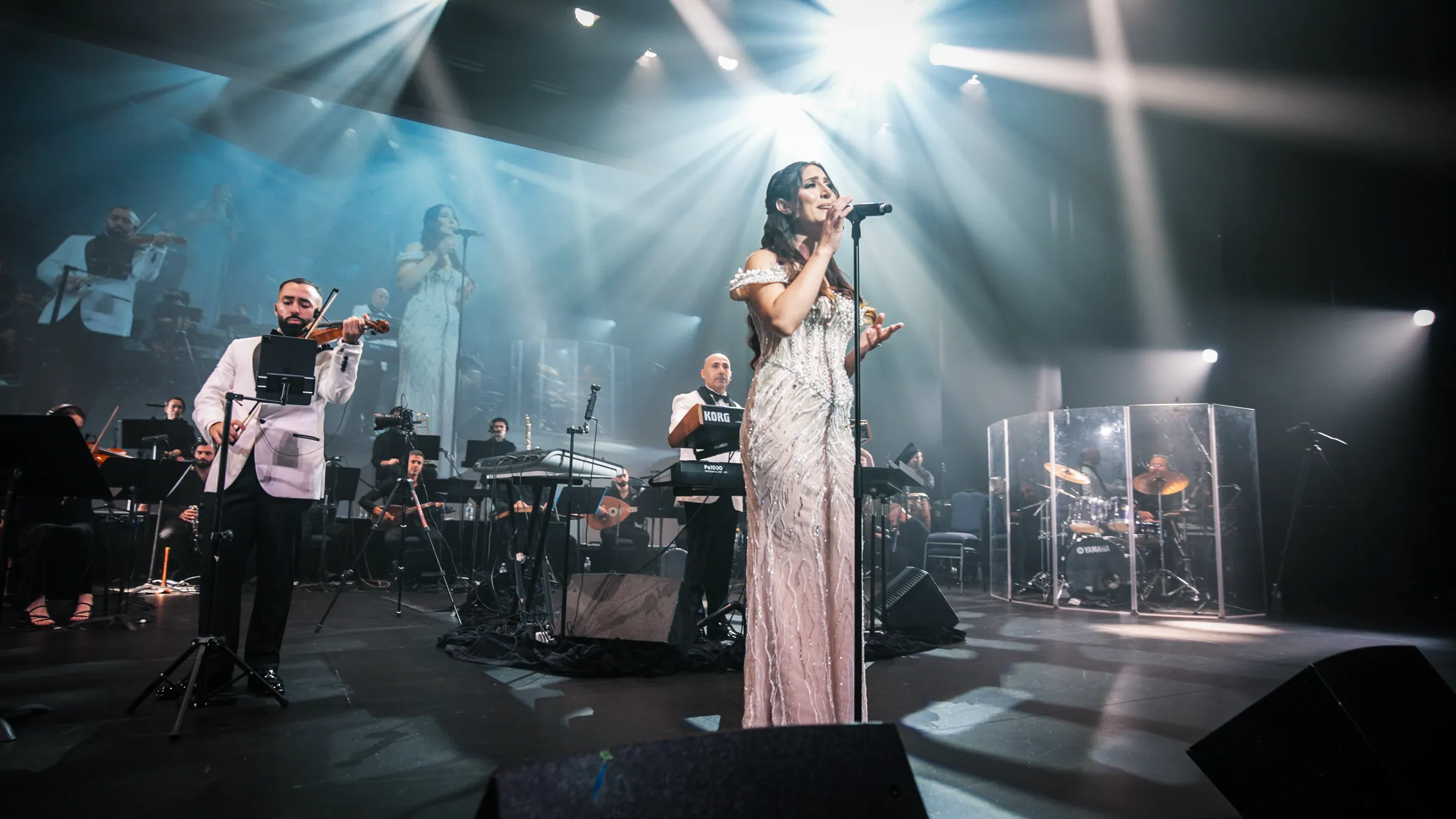 A vocalist in white with Jad on violin and the orchestra, under blue beams