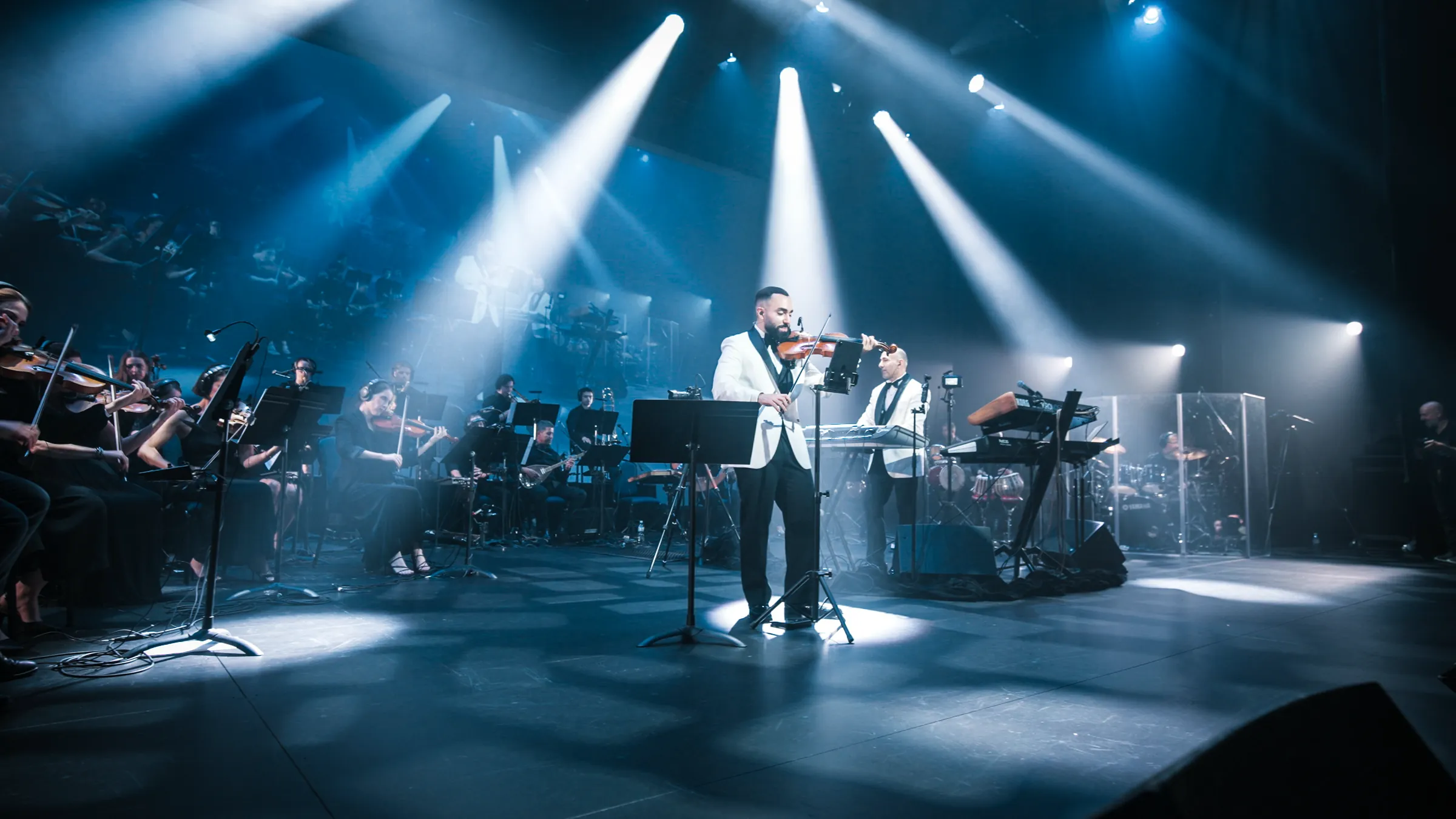 The Jad & Rabih orchestra on stage in 2026, under crossing white beams of light